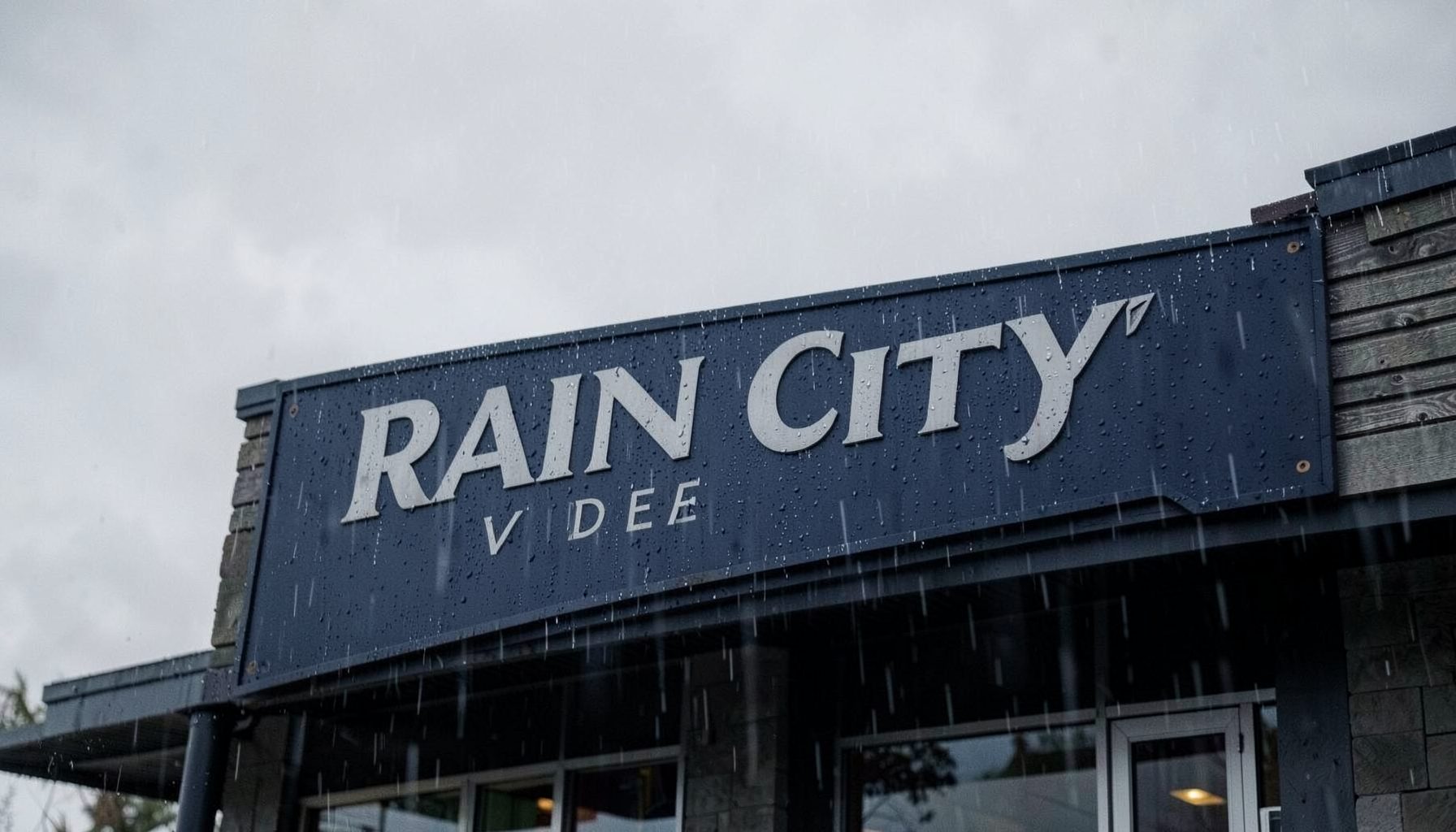 Rain City Video logo — a moody, rain-speckled storefront sign in deep navy and muted silver tones against a soft gray Seattle backdrop